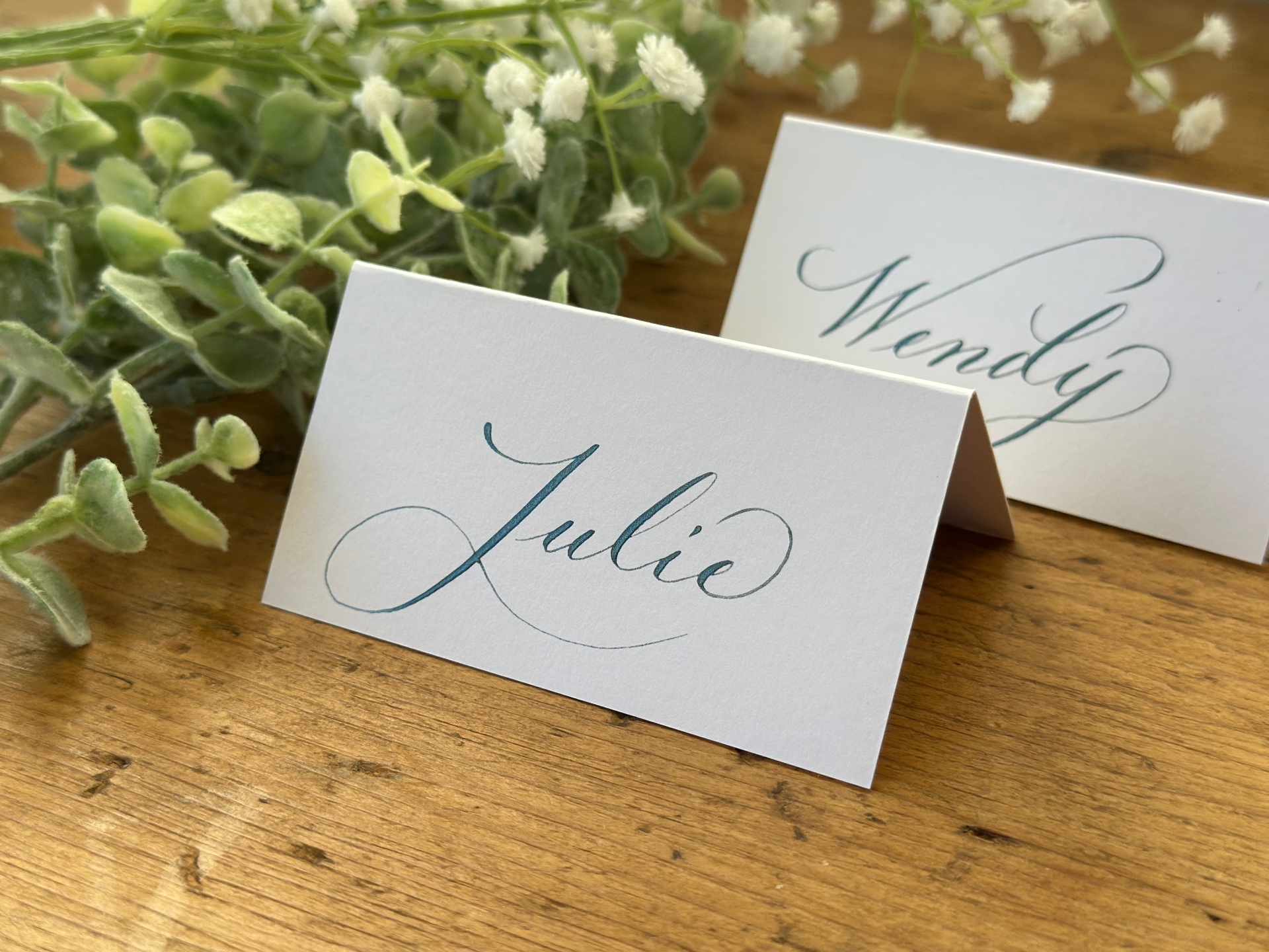 Wedding place cards handwritten in teal ink, beside eucalyptus